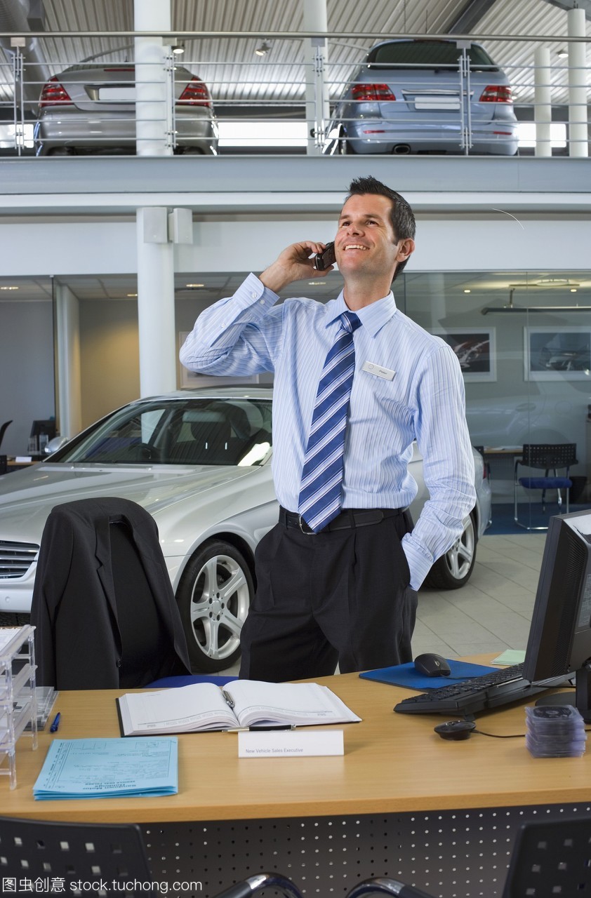 汽車銷售員站在柜臺旁的大型汽車陳列室里,用手機微笑著